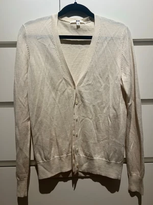 Beige tunn kofta från Uniqlo - Säljer en stilren beige kofta från Uniqlo med v-ringning och knäppning framtill. Koftan är tunnstickad med långa ärmar och ribbade muddar vid ärmslut och nederkant. Perfekt att slänga över en t-shirt eller linne för en enkel look. Väldigt snygg. Storlek m men passar xs ocksp
