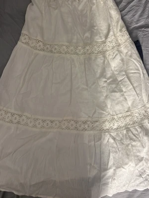 Vit långkjol med spetsdetaljer - Romantisk vit långkjol med breda spetsband och lätt rynkad midja. Kjolen har en luftig och bohemisk känsla, perfekt för varma dagar. Tillverkad i ett mjukt och svalt tyg som ger en avslappnad look.