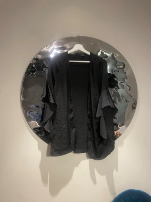 Svart kimono med volangärmar - Säljer en svart kimono-inspirerad cardigan med vida volangärmar. Materialet är tunt och har en lätt glansig finish som ger ett snyggt fall. Perfekt att slänga över en topp för en cool och avslappnad look.