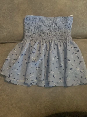 Ljusblå blommig kortkjol - Supersöt ljusblå kortkjol med små vita och blå blommor. Kjolen har smock i midjan för en bekväm passform och ett lätt, luftigt tyg som passar perfekt till varmare dagar. Från Gina trocot 