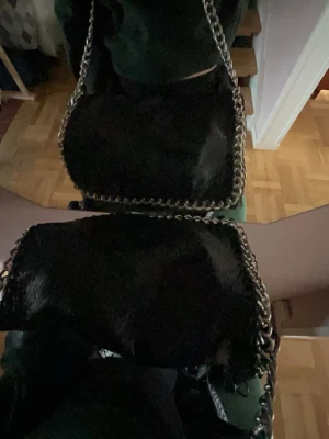 Svart axelväska med kedjedetaljer - Snygg svart axelväska från Tiamo med grova silverfärgade kedjor längs kanterna och som axelrem. Väskan har en mjuk, något glansig yta i konstläder och en fyrkantig form. Perfekt för dig som gillar en edgy och trendig stil. Lite avskavd färg på de 2 öglorna tyvärr. 💗