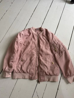 Ljusrosa bomberjacka med gulddetaljer - Snygg ljusrosa bomberjacka med dragkedja framtill och guldfärgade knappar på fickorna. Jackan har ribbade muddar vid ärmslut och nederkant samt en liten ficka med dragkedja på ena ärmen. Perfekt för dig som vill ha en stilren och trendig look.