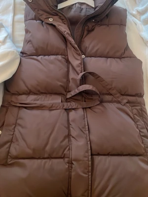 Brun lång dunväst med bälte - Säljer en snygg brun lång dunväst med bälte i midjan och hög krage. Västen har quiltad design och stängs med både dragkedja och tryckknappar. Perfekt för lager på lager och ger en trendig siluett. Passar dig som vill ha både stil och värme.