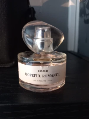 Hopeful Romantic Eau de Toilette - Säljer en elegant parfymflaska från Hopeful Romantic med en unik, asymmetrisk transparent kork. Flaskan är rund och gjord av glas med ljusrosa doftvätska. Märkt som vegan och Eau de Toilette. Perfekt för dig som gillar stilrena och moderna dofter. Andvänd ca 1/3.