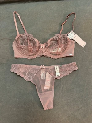 Rosa spets-set från Lindex - Sött underklädesset från Lindex / Ella M i ljusrosa / dammrosa spets med blommigt mönster. Setet består av en bygel-bh med justerbara axelband och en matchande trosa, båda dekorerade med små rosetter och pärldetaljer framtill. Materialet är genomskinlig mesh och spets för en romantisk look🌸Självklart helt oanvända båda två! 