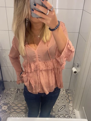 Ljusrosa volangblus med v-ringning - Söt ljusrosa blus med v-ringning och volangdetaljer. Blusen har långa ärmar och tunna knytband vid halsen. Tillverkad i ett lätt, luftigt tyg som ger en avslappnad och romantisk look. Perfekt att matcha med jeans eller kjol.