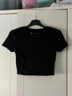 Svart croppad ribbad topp - En svart croppad topp med ribbad struktur och korta ärmar. Enkel och stilren design som passar perfekt till jeans eller kjol. Rund halsringning och figurnära passform.