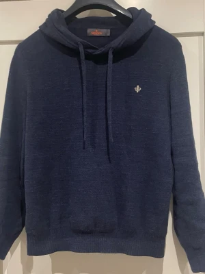 Mörkblå hoodie från Morris - Mörkblå hoodie från Morris med broderad guldfärgad lilja på bröstet. Tröjan har dragsko i huvan och är tillverkad i ett mjukt, stickat material. Perfekt för en avslappnad och stilren look.