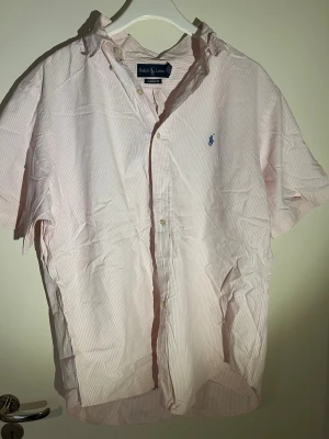 Rosa- och vitrandig skjorta Ralph Lauren - Kortärmad skjorta från Ralph Lauren i ljusrosa och vitt med tunna ränder. Klassisk krage, knappar framtill och den ikoniska blå loggan broderad på bröstet. Tillverkad i bomull för en sopren och luftig känsla.