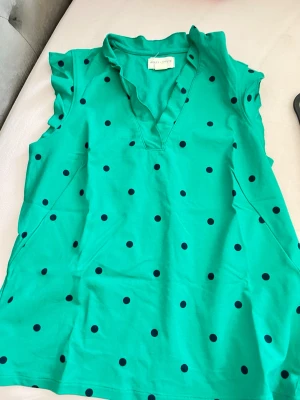 Grön prickig topp Holly & Whyte - Snygg grön topp från Holly & Whyte by Lindex med mörkblå prickar. Toppen har volangdetaljer vid ärmöppningarna, v-ringning och är är warmlös. Perfekt för dig som gillar färg och solid mönster.