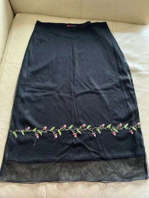Svart kjol med broderade blommor - Svart kjol med broderade blommor i rosa och grönt längs nederkanten. Kjolen har en rak passform och är tillverkad i ett tunt, lätt material med transparent kant längst ner. Perfekt för dig som gillar unika detaljer.