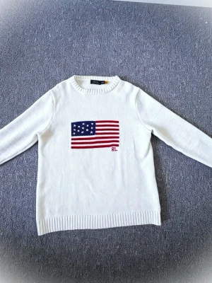 Vit stickad tröja Ralph Lauren - Vit stickad tröja från Ralph Lauren med amerikansk flagga broderad på bröstet och RL-logga i rött. Tröjan har rund halsringning, långa ärmar och ribbade muddar vid ärmslut och nederkant. Perfekt för dig som gillar klassisk och stilren design.