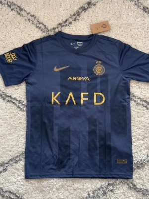 Blå Al Nassr Ronaldo matchtröja Nike - Snygg mörkblå Al Nassr matchtröja från Nike med guldiga detaljer och tryck. Framsidan har klubbmärke, sponsorloggor och Nike-logga, medan baksidan har 'Ronaldo' och nummer 7 i guld. Tillverkad i lätt och ventilerande material, perfekt för fotboll eller träning.