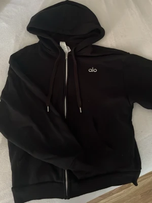Svart zipue hoodie från Alo - Säljer en svart hoodie från Alo med dragkedja framtill och justerbar huva med snören. Diskret Alo-logga på bröstet. Hoodien har ribbade muddar och är tillverkad i mjukt material som känns skönt mot huden. Perfekt för en sopren och chill look.