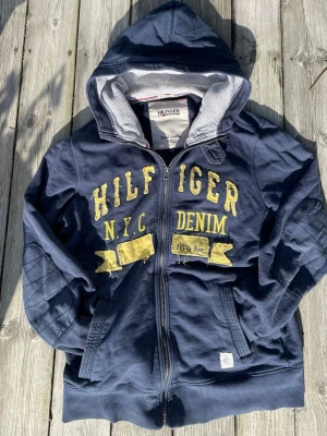 Tommy hilfiger Class of ’85 Hoodie  - Tommy Hilfiger Denim “Class of ’85” zip hoodie i mörkblått. Inspirerad av märkets klassiska college-estetik från 80-talet. Stor broderad Hilfiger-logga över bröstet med N.Y.C och denim-patchar framtill. Distressed detaljer i loggan ger en riktigt bra vintage look.  Tjock och bekväm hoodie med zip, fickor fram och hög krage. Perfekt för Y2K / vintage / collegiate style.