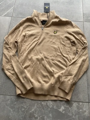 Beige stickad tröja från Lyle & Scott - Stickad beige tröja från Lyle & Scott med halv dragkedja och deras klassiska gula logga på bröstet. Tröjan är helt ny och aldrig använd.