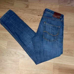 Blå jeans med slitningar - Snygga blå jeans från Jack & Jones med en cool slitning på framsidan. De har en klassisk femficksdesign, jeansen är i bra skick och inga tecken på användning. Perfekta för en avslappnad stil. Nypris 899kr
