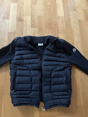 Svart cardiganjacka från Moncler - Snygg svart cardiganjacka från Moncler med vadderad front och stickade ärmar. Jackan har dragkedja framtill, två fickor med dragkedja och Moncler-logga på ärmen. Perfekt för lager-på-lager och streetstyle. Materialmix av dun och ull ger både värme och stil.