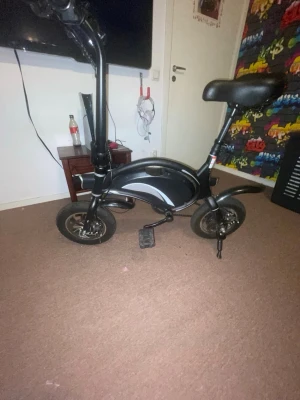 Svart elcykel med kompakt design - Säljer en svart elcykel med modern och kompakt design. Cykeln har små hjul, ett robust ram och ett bekvämt sadel. Perfekt för kortare resor och enkel att förvara. Pedaler och stöd ingår, och cykeln har en stilren look.