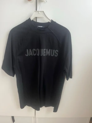 Svart Jacquemus t-shirt - Snygg svart t-shirt från Jacquemus med stor logga tryckt framtill. Klassisk rak passform och rund halsringning. Tillverkad i mjukt bomullsmaterial som känns skönt mot huden. Perfekt för en clean och trendig look. Nypriset var 3000 kr. Köpt på Minto. Modellen är oversize. Skicket är 9/10 endast använd 3 gånger 