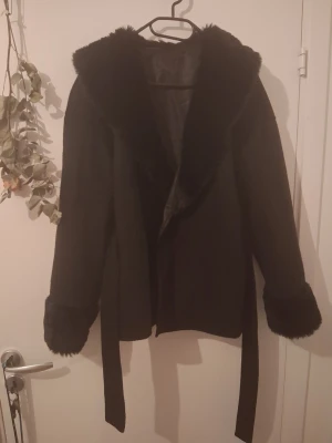 Svart kappa med fuskpälsdetaljer - Stilren svart kappa med bred krage och muddar i mjuk fuskpäls. Kappan har ett knytband i midjan för en snygg siluett och är helsvart, vilket gör den lätt att matcha. Perfekt för kyliga dagar när du vill vara både varm och trendig.