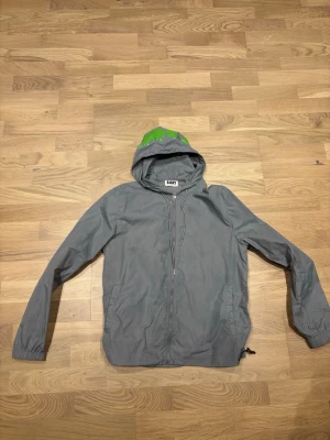 Grå vindjacka från Risky med huva - Snygg grå vindjacka från Risky med dragkedja framtill och justerbar huva. Jackan har en cool grön detalj på huvan och elastiska muddar vid ärmsluten. Tillverkad i lätt och vindtätt material, perfekt för blåsiga dagar. Ända defekten är att fickan har fått små fläckar när jag skadade mig går antagligen att tvätta men har inte provat 