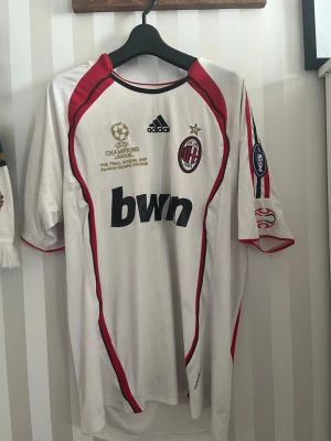 AC Milan bortatröja Kaka 22 Adidas - Säljer en vit AC Milan bortatröja från Adidas med röda och svarta detaljer, Champions League-märke och tryck Kaka 22 på ryggen. Tröjan har klubbmärke, sponsortryck och officiella patches. Perfekt för fotbollsfans och samlare.