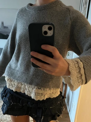 Grå stickad tröja med spetsdetaljer - Mysig grå stickad tröja med rund hals och dekorativa beige spetsdetaljer vid ärmslut och nederkant. Tröjan har en avslappnad passform och långa ärmar. Perfekt för lager på lager och en trendig vardagslook.