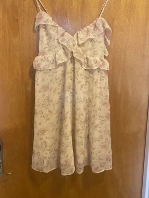 Blommig kortklänning med volang - Söt kortklänning med tunna axelband och volangdetaljer framtill och baktill. Klänningen har ett ljust blommigt mönster i pastellfärger och knyt i ryggen. Perfekt för varma dagar och har en luftig, flowy passform.