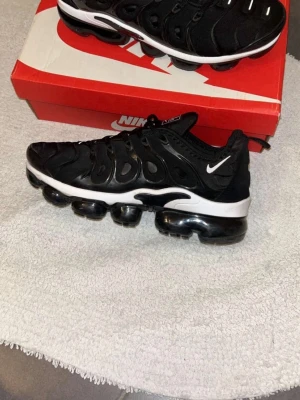 Nike Air Vapormax Plus svarta sneakers - Nike Air Vapormax Plus sneakers i svart med vit sula och små vita detaljer. Ovandelen är i textil och syntet med en unik burdesign och synlig Air-dämpning i sulan. Swoosh-logga på sidan och klassisk Vapormax-bubbelsula för extra komfort.