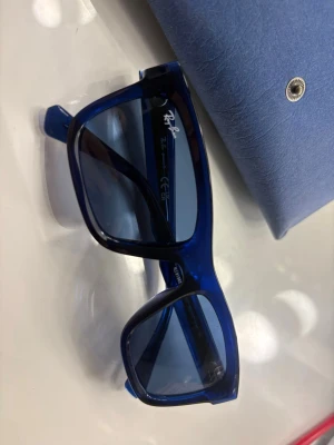 Blå Ray-Ban Junior solglasögon - Snygga blå Ray-Ban Junior solglasögon med rektangulär form och transparent båge. Linsfärgen är mörkblå och bågarna är tillverkade i syntetmaterial. Perfekt för dig som vill ha ett stilrent och ungdomligt accessoar.