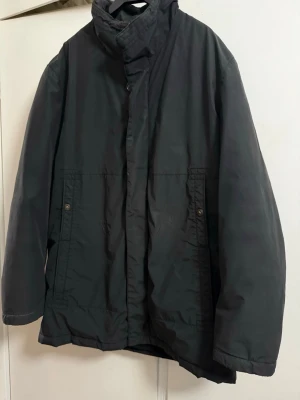 Svart vindjacka med hög krage från Boss - Stilren svart vindjacka med hög krage och dold dragkedja framtill. Jackan har två framfickor med knappstängning och en rak passform. Materialet är slätt och vattenavvisande, perfekt för blåsiga dagar. Enkel design utan synligt märke.