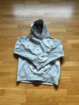 Grå hoodie denim tears - Säljer en grå hoodie med stora vita moln sopsäcksmönster över hela tröjan. Hoodien har en klassisk huva och magficka framtill. Perfekt för dig som gillar streetwear och vill sticka ut lite extra. Mjuk och skön bomullsblandning.