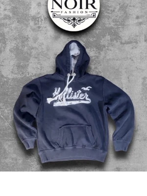 Blå Hollister hoodie och vit 2 st - Mörkblå hoodie från Hollister med vit broderad logga och fågel på bröstet. Klassisk känguruficka, ribbade muddar och vit dragsko i huvan. Perfekt för en avslappnad streetwear-look. 2 st fläckfria 