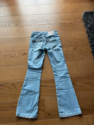Ljusblå bootcut jeans från True Religion - Säljer ett par ljusblå bootcut jeans från True Religion med klassiska kontrastsömmar och fickor med lock och knappar bak. Jeansen banar ut nedtill för en snygg retrostil och adjusting passform. Perfekta för dig som gillar en avslappnad men trendig look. Dom är upplagda och passar längd: 155-160 cm. Stl 23