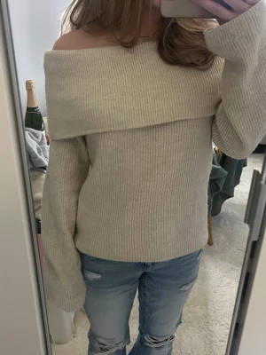Beige off shoulder stickad tröja - Säljer en beige ribbstickad off shoulder-tröja med bred krage. Tröjan har långa ärmar och en avslappnad passform, perfekt för en trendig och mysig look. Materialet ser mjukt och bekvämt ut, perfekt att styla med jeans.