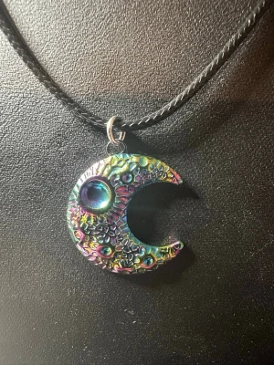Regnbågsskimmrande månformad halsband - Unikt halsband med ett månformat hänge i metall som skimrar i regnbågens färger. Hänget har en cool struktur med små upphöjda detaljer och en större pärlliknande dekoration. Hängs på ett svart, band som ger en edgy vibe.