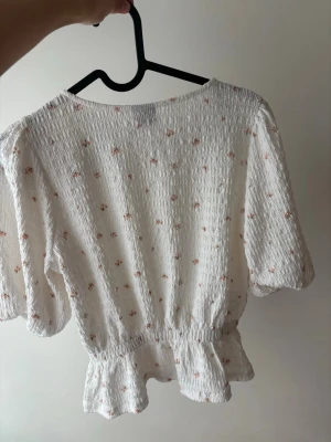 Vit blommig blus från Monki - Supersöt vit blus från Monki med små blommor i rosa toner. Blusen har korta puffärmar, smockat tyg och volangkant nedtill. Perfekt för en somrig och romantisk look. Materialet känns lätt och luftigt.