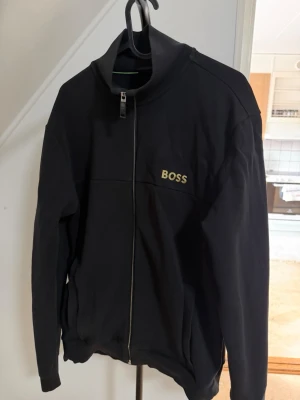 Svart zip hoodie från BOSS - Snygg svart hoodie från BOSS med dragkedja framtill och guldfärgad logga på bröstet. Hoodien har hög krage och ribbade muddar vid ärmar och nederkant. Perfekt för en clean och stilren look.