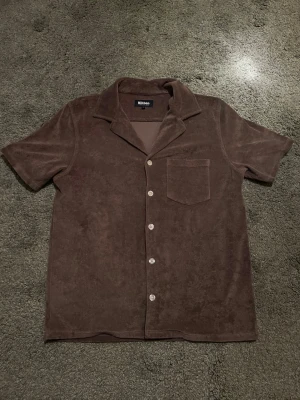 Brun frottéskjorta från Nikben - Brun skjorta från Nikben i mjuk frotté med kort ärm, klassisk krage och knäppning framtill. Skjortan har en bröstficka och avslappnad passform, perfekt för en chill och trendig look.