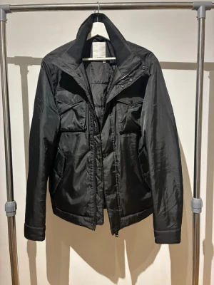 Svart vindjacka med fickor från J.Lindberg - Stilren svart vindjacka med flera praktiska fickor framtill och hög krage. Jackan har dragkedja och tryckknappar, samt en lätt glansig finish. Perfekt för dig som vill ha en enkel och funktionell jacka med modern look.  Jackan är använd ytterst få gånger och är i otroligt bra skick.                                                    Vid intresse skriv i DM🙌🏽