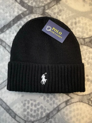 Svart ribbstickad mössa Polo Ralph Lauren - Säljer en svart ribbstickad mössa från Polo Ralph Lauren med den klassiska vita broderade loggan framtill. Mössan har uppvikt kant och är tillverkad i mjukt material som håller dig varm. Perfekt accessoar för kalla dagar.