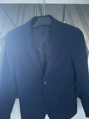 Mörkblå klassisk kavaj - Stilren mörkblå kavaj med dubbelknäppning och klassisk krage. Kavajen har två framfickor och en bröstficka, samt är helfodrad för en snygg siluett. Perfekt för dig som vill ha en tidlös och elegant look.