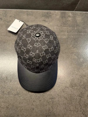 Svart Gucci keps med GG-mönster - Storlek M Snygg svart keps från Gucci med klassiskt GG-monogram i grått över hela ovandelen. Skärmen är i svart läder-look och kepsen har en rundad form. Perfekt för dig som vill ha en exklusiv accessoar med streetwear-vibe.