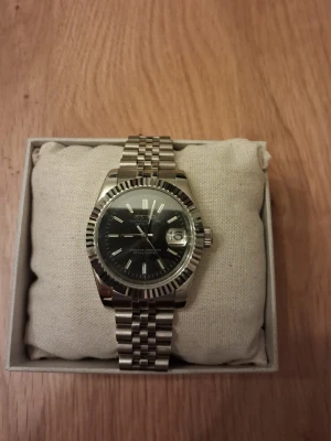 Seiko Klocka - Seiko armbandsur med automatisk urverk. Klockan har en svart urtavla och ett datumfönster. Boetten och armbandet är tillverkade av stål. Extra länkbitar, putsduk och ask medföljer.