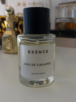 ESSNCE Eau de Caramel parfym - ESSNCE Eau de Caramel är en eau de parfum med söt karamell-doft. Flaskan är rund och genomskinlig med svart matt kork och vit etikett. Parfymen har en ljusgul vätska och stilren design som passar perfekt på parfymhyllan.
