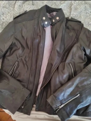 Petroff Svart skinnjacka med ståkrage - Snygg svart skinnjacka med ståkrage och tryckknappar. Jackan har dragkedjor på ärmarna och framfickor samt axelklaffar. Insidan är fodrad med randigt tyg. Perfekt för en cool och stilren look. Storlek XL