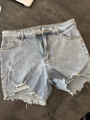 Ljusblå denimshorts med slitningar - Snygga ljusblå jeansshorts i denim med råa kanter och slitna detaljer framtill. Klassisk femficksmodell med bälteshällor och knappstängning. Perfekta för sommaren och ger en avslappnad streetstyle-look.