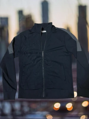 Svart sportjacka med grå detaljer - Svart sportjacka med hel dragkedja framtill och gråa paneler på axlarna. Jackan har ståkrage, långa ärmar och två sidofickor. Perfekt för träning eller chill dagar. Materialet känns mjukt och följsamt.