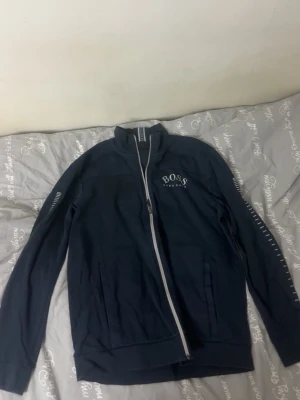 Mörk Blå ziptröja från BOSS - Snygg Mörk blå ziptröja från BOSS med vit dragkedja och logga på bröstet. Tröjan har hög krage, långa ärmar och ribbade muddar. Perfekt för en stilren och sportig look. Materialet känns mjukt och bekvämt. Finns lite skador för mer info kontakta mig